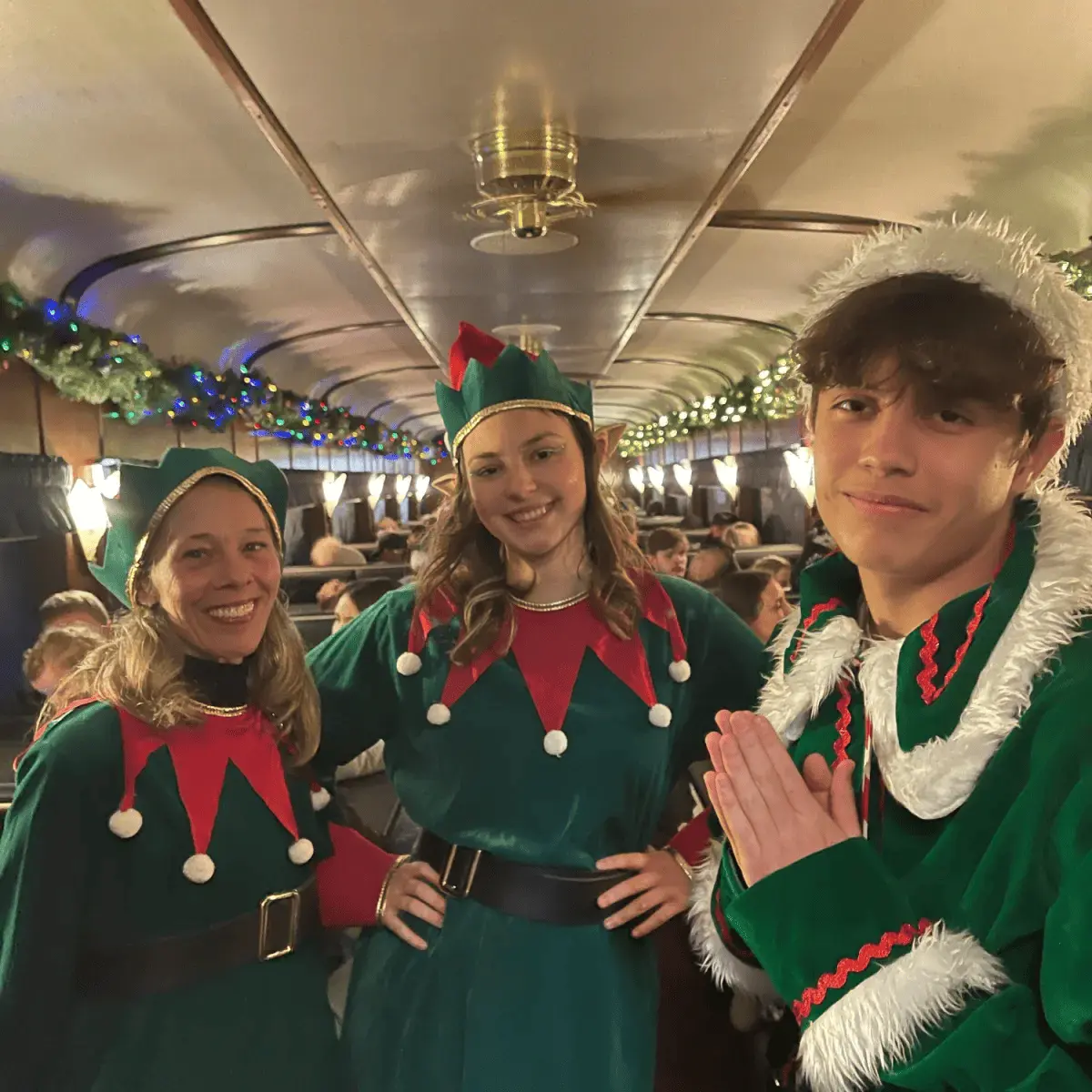 Three people in elf costumes smiling inside a decorated train car.