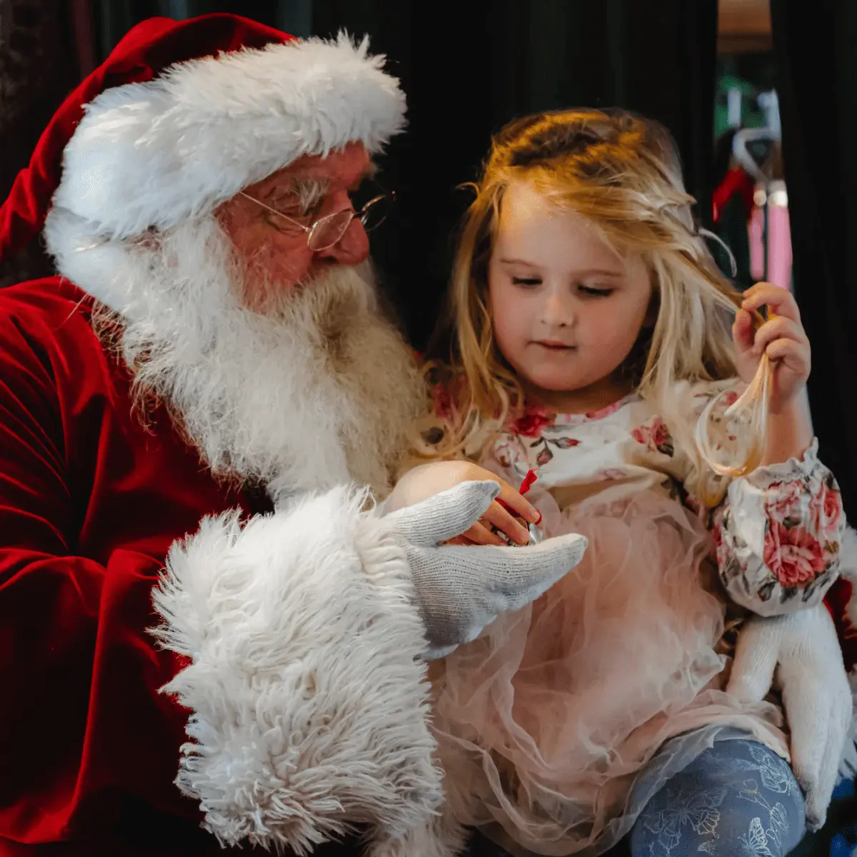 Santa Claus holding a young girl on his lap, both looking at a small gift in her hand.