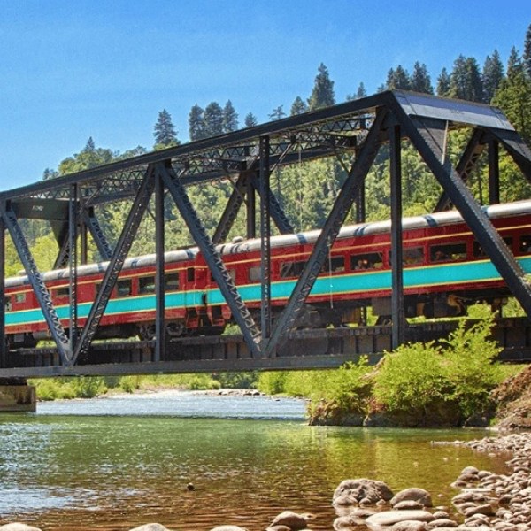 Train crosses a truss bridge over a river, surrounded by trees and blue sky.