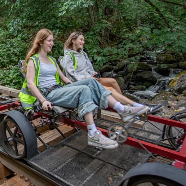 Two women pedaling a handcar on railway tracks in a forested area.