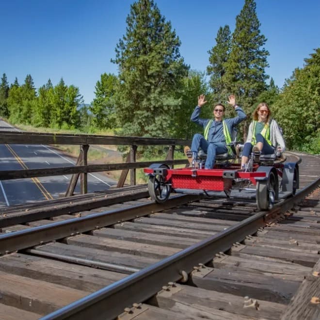 Two people ride a rail bike on tracks with trees and a road nearby.