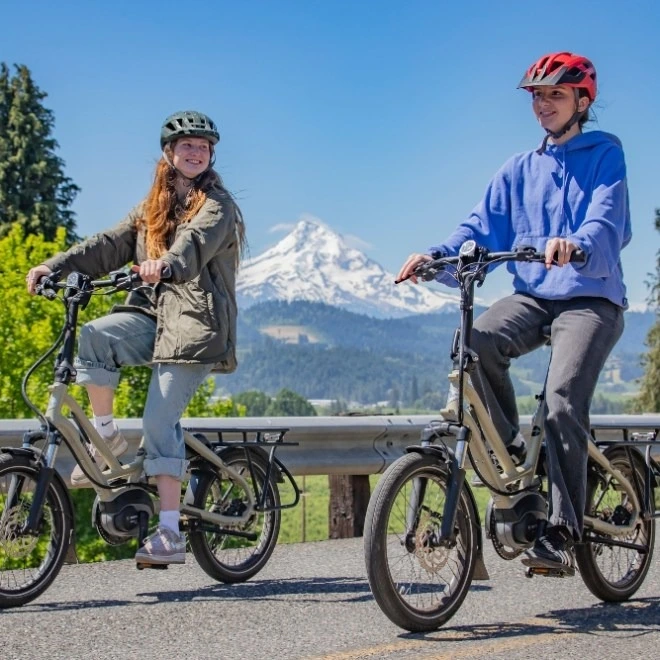 Two people wearing helmets ride e-bikes with a snowy mountain in the background.