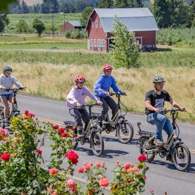 Four people riding e-bikes on a road with a red barn and fields in the background.