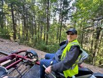 Man in safety vest smiles while seated on a track vehicle in a forested area.